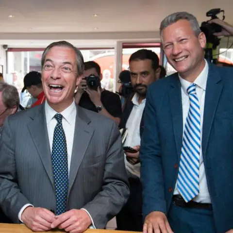 Getty Images Nigel Farage, wearing a grey suit and blue and grey tie, is laughing with Nathan Gill, a tall man wearing a navy blue suit and a blue and light grey stripy tie, is next to him also laughing. Both are at a press event and standing in front of a table. 