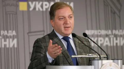 UNIAN A brown haired man with a grey check suit wearing a light blue shirt and a navy blue tie in front of two black microphones giving a speech in front of a Russian sign 