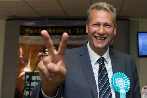 Getty Images A white brown-haired politician, wearing a blue suit, white shirt and stripy blue, white and grey tie, is smiling at the camera at an election count while putting a victory sign up to the camera lens. He is wearing a light blue Brexit Party rosette. 