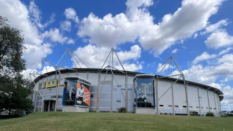 An exterior view of a modern sports stadium with a curved silver facade and triangular support structures on the roof. Large banners are displayed on the front, and the stadium is surrounded by a grassed area.