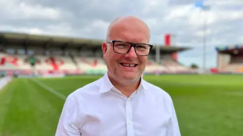 BBC A man wearing a white button-up shirt standing on the edge of a green sports field, with a stadium in the background featuring red and white seating.