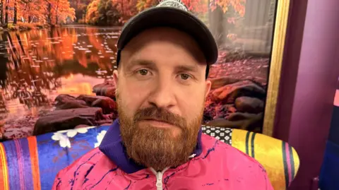 BBC/Jasmine Lowe A man with brown eyes and a ginger beard looking into the camera. He has a cap on and has bushy eyebrows. He is sitting in a colourful room where he films and records his podcasts wearing a pink and purple zip-up jacket. 