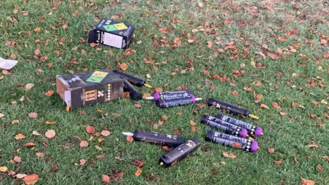 BBC News A patch of autumnal grass with scattered brown leaves resting on it. There are also about a dozen large canisters with danger warnings printed all over the packaging. Two boxes are also littered with small balloons weaved into the grass. 