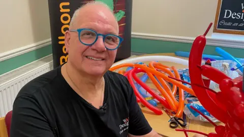 BBC A man in his sixties smiles at the camera. He is sitting next to a table which is covered in modelling balloons and other equipment, including scissors. There are several red balloons in the foreground, which make up the back of one of his dresses. He is wearing bright blue glasses.