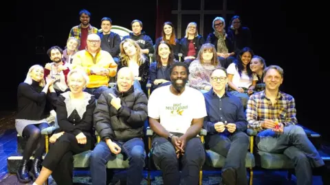 Kofi Smiles A group of 21 people seated in three rows on a small stage. The seating is arranged in tiers. The lighting is bright on the group, in comparison to the pitch-black background. There is a mixture of males and females on each row, some lean into each other smiling at the camera, others hold small toys in their hand and some point at each other. 