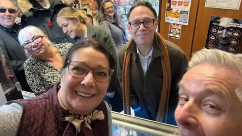 Dinsdale's A group selfie with a mixture of males and females spread throughout in a small joke shop. A brunette female with large framed glasses and a burgundy gillete takes the photo and smiles. Behind her there are two males and three females who are leaning to and away from the camera, their heads are partially or fully cut out of the frame. To the right is a dark-haired man with black glasses, a khaki coat and mustard coloured scarf - he leans towards the camera. A white-haired man is partially cut out of frame, he has his mouth open wide.