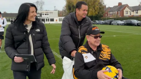 BBC/Jasmine Lowe Hull City owner Acun Ilicali, a man in long black coat, pushes Owen Griffiths, an elderly man wearing a Hull City tracksuit and holding a yellow football, in his wheelchair. A woman with dark hair and wearing a black jacket smiles as she walks beside them. 
