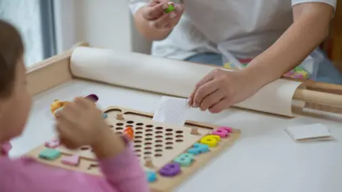 Getty Images A stock photo of a blurred child in a long pink top who can be seen holding up their hand and looking at wooden boards with holes in it and colourful numbers down the side and shapes. An older person is holding a paper with circles on it at the front of the wooden board and is also holding something green and has a plastic box rested in their lap.