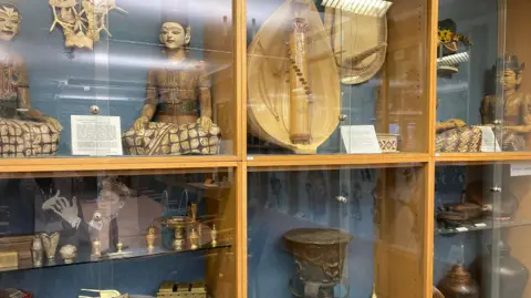 An array of South East Asian artefacts in a wall-mounted display case. The items include figurines, and pots.