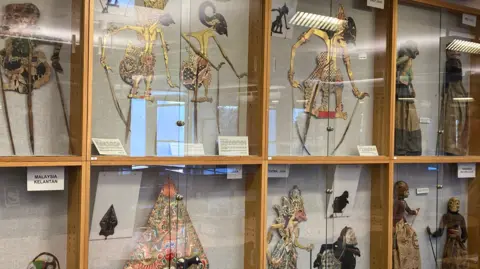 Colourful figurines, including some from Indonesia, in a glass display case. The individual displays are separated by wooden frames.