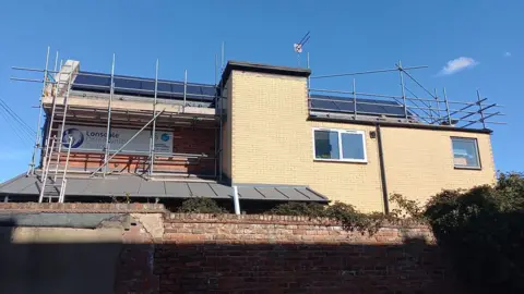 Power Hull The solar panels being installed on the roof of the Lonsdale Community Centre a yellow brick building with a wall and hedges at the front. The building is surrounded by scaffolding.
