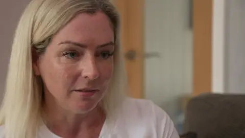 BBC A woman in her 40s has long blonde hair swept back behind her ears and wears a white T-shirt. She is sitting in a living room with a pensive look on her face. Wood and glass doors and a grey sofa can be seen in the background, which is out of focus.