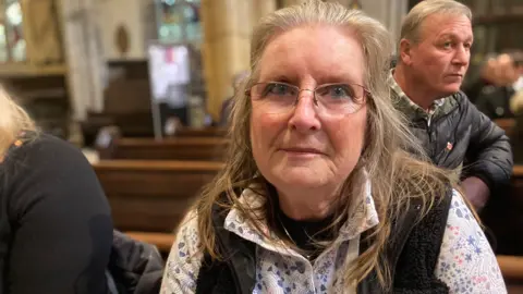 BBC Ms Mooring wears glasses and looks into the camera from where she is sitting in the pew.
