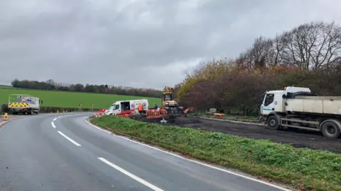 Paul Hopton A road which has no cars driving along it bends round to the right. On the right a grass verge can be seen with maintenance vehicles and a digger which has taken up some of the grass to leave a neat line of soil. Cones can also be seen dotted around the road.