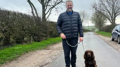 Handout Councillor Paul Hopton - a man with short grey hair smiling with a navy coat standing along a single-access lane with his dog sitting by his side on a lead. The dog is a brown cocker spaniel.