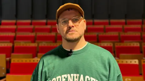 A head and shoulders shot of Paul Smith looking at the camera. He is wearing glasses, an orange cap and a green sweater with "Copenhagen" in white bold capital letters printed on the front. He is stood in front of red theatre chairs. The walls are black.