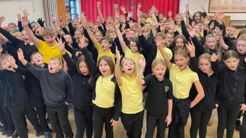 BBC/Simon Thake A large group of schoolchildren in black uniforms with yellow polo shirts all have one hand in the air and are looking towards the camera. Their faces show they are singing. They are indoors in a hall with red curtains in the background and a noticeboard that says British Values on it.