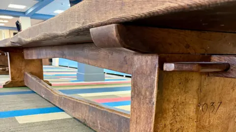 Hawleys Auctioneers & Valuers Close-up of a long, heavy wooden table with thick legs and a horizontal support beam, placed on a carpet with multicoloured stripes. The wood shows signs of age and wear, and the year ‘1937’ is carved into one of the legs.