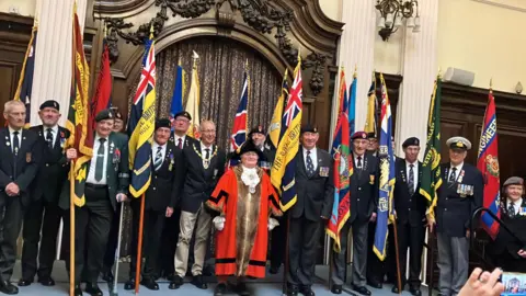 BBC/Naj Modak A group of 14 smartly dressed men and a woman, holding Royal British Legion flags and other standards, stand in a line in a grand hall. They wear regimental caps and ties and some have medals. In front of them is a woman dressed in red robes and a black admiral's hat. 