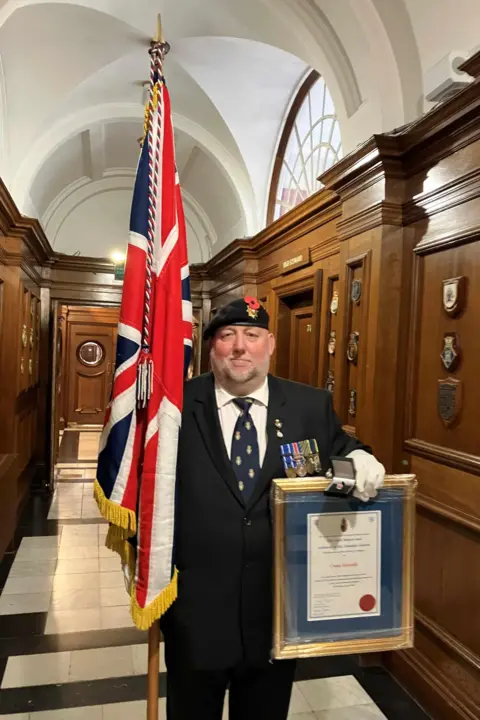 BBC/Naj Modak Man in smart blazer and regimental beret and tie holding a medal and a framed certificate in his left hand and a Union flag standard in the other. He is standing in a wood-panelled corridor.