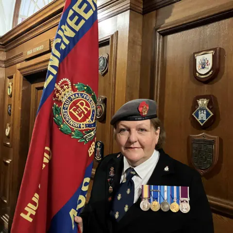BBC/Naj Modak A woman wearing a black blazer, regimental beret, tie and a row of medals stands in a grand-looking, wood-panelled corridor while holding the flag of a regimental association, which is coloured scarlet and blue and includes a regimental badge.