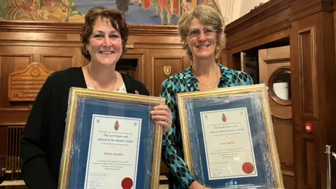 BBC/Naj Modak Two award recipients - a woman with brown hair and a woman with blonde hair - standing indoors holding framed certificates with blue backgrounds and gold borders. The certificates feature printed text and a red wax seal. Behind them is a wooden-panelled wall with a shield emblem and a large mural above.