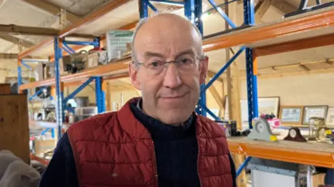 BBC Rob Byass. A 69-year-old man with glasses wearing a blue turtle neck and red puffer gilet. He is standing in a barn. To his left is a shelf holding several donated clocks. 