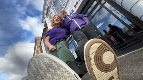 A low-angle view of Frances Finn on the left and standing outside the BBC Radio Lincolnshire studio with large glass windows and a white facade. The woman on the left is wearing green trousers and a purple top, while the man on the right wears dark trousers and a purple jacket.
