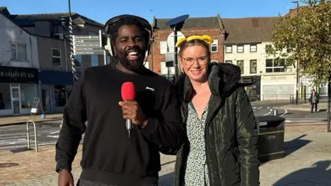 BBC Kofi Smiles and Carla Greene stand on a street and smile into the camera. Kofi, on the left, is wearing a black jumper, headphones and holding a microphone. Carla, on the right, is wearing yellow bear ears and glasses. A number of shops and trees are visible in the background.
