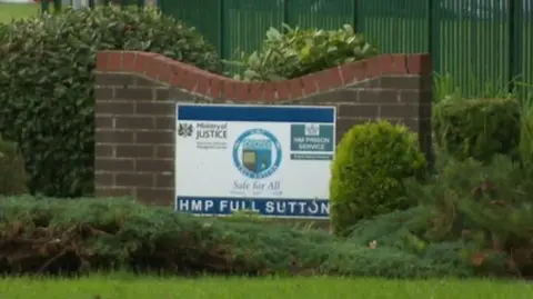 BBC A sign reading HMP Full Sutton on a small brick wall surrounded by bushes.