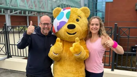 Pudsey Bear, a large yellow bear mascot with a colourful polka-dotted bandage over one eye, standing outdoors in front of a metal railing and a modern building with glass panels and a red brick wall. The mascot is giving two thumbs up and is flanked by two people on either side, both also giving thumbs up. 