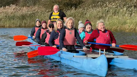 Laughing and smiling 10 women sit side by side in a blue bell boat. Pete Cogley from Paddlers for Life stands at the helm behind them wearing a yellow life vest. The women are all wearing red life vests and holding red oars. Pete has grey hair and a beard and is waring a black t shirt with a yellow life vest. 