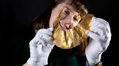 North Yorkshire Council A person is holding a delicate, golden, fan-shaped object called a lunula with both hands. They are wearing clean white gloves. The object shines under the light, with a textured surface. The person holding the lunula is dressed in a dark green top and wearing a blue wristwatch on their left wrist. The background is dark, which makes the golden object and the gloved hands stand out clearly. 