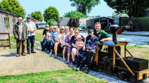 JAMES ARDIN A mixed group of adults and children are sitting on the carriage of a miniature, ride-on train, behind an old-fashioned engine made of wood and black metal. The railway line can be seen snaking around a large garden, with shed-type buildings in the background. Two men are standing on wooden decking to the left. It is a sunny day, with a blue sky and green trees in the background.
