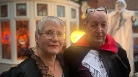 Simon Thake/BBC An elderly man and women pose together in black fancy dress costumes. She has short hair and glasses. He is wearing a dracula outfit. Behind them a house has been decorated with ghosts and pumpkins