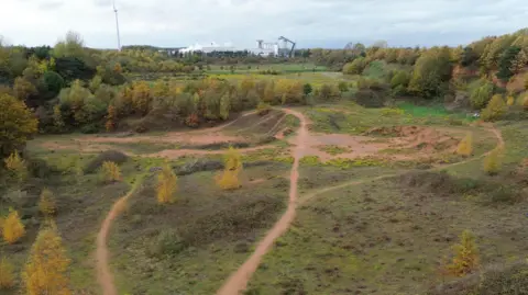 Footage from a drone shows the former quarry site. There overgrown scrubland surrounded by young trees. There is an industrial area in the background. 