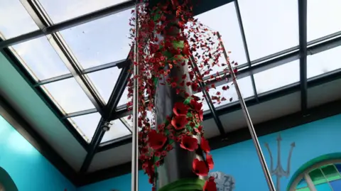 The picture shows a black pole running down the middle. It's adorned with red poppies of different sizes. Above is is a glass roof. 