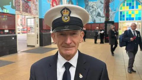 The photo shows a man looking at the camera. He's wearing a military suit and tie, and a hat. He's standing in front of a train station. There are poppy displays in the background.