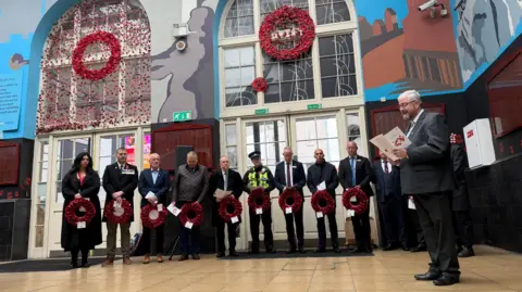 BBC The picture shows a row of people all holding wreaths covered in poppies. They're standing in a train station, and are all looking at a man to the right hand side of them, who is reading an order of service. 
