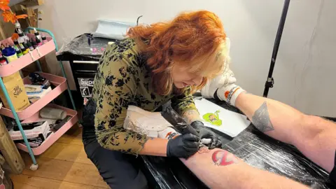 BBC/Becki Bowden Tattoo artist Holly Dosdale at work. She is working on a tattoo on a man's leg. 