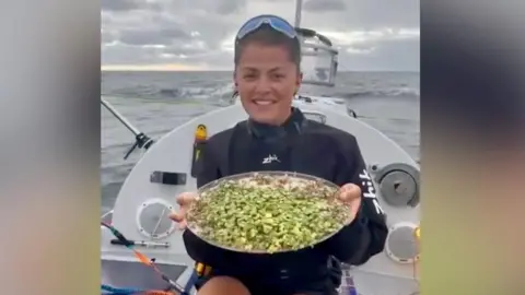Seas the Day Miriam Payne is sitting on her rowing boat in the Pacific Ocean. She is holding a circular tray of greens. She is wearing shorts and a black top and has sunglasses on her head.