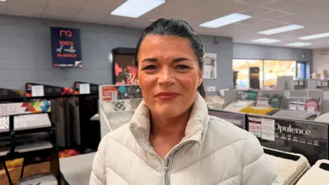 BBC News/Jasmine Lowe A lady in a white jacket in a carpet shop, with black hair and makeup on. She is the manager at the store 