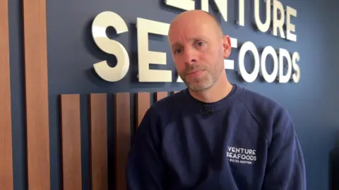 BBC A man with a bald head is standing in his office in front of a wall which shows the business logo of Venture Seafoods. He is wearing a navy blue jumper, which also has the business logo printed on the left side. He's looking pensive as he speaks to camera.