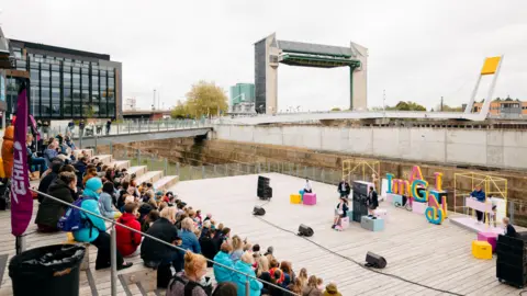 Tom Arran Crowds of people sat on steps watching a play on a wooden stage by a river in Hull. The sky is overcast.