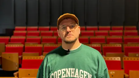 BBC A head and shoulders shot of Paul Smith looking at the camera. He is wearing glasses, an orange cap and a green sweater with "Copenhagen" in white bold capital letters printed on the front. He is stood in front of red theatre chairs. The walls are black.