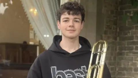 East Riding of Yorkshire Council A young man with dark wavy hair, wearing a hooded top and jeans, stands with a trombone. He is smiling. 