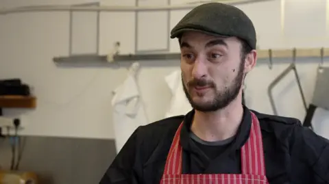 BBC A butcher stands in his shop, which has a white a grey wall with hooks for white aprons. He has a dark brown beard and is wearing black overalls, a black cap and a red-and-white striped butcher's apron. 