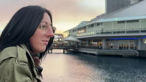 BBC News/Jasmine Lowe A lady looking across water, the sky is light pink and she has black hair. She's wearing clear glasses and a green coat. 