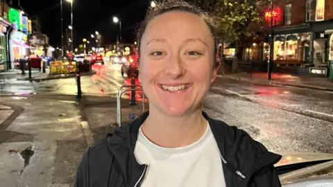 A woman dark hair tied back standing on the pavement next to a street in Hull. In the background is a row of shops and bars. Claire is wearing a white t-shirt and has an unzipped black running jacket on.