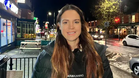 BBC Sophie stands in a busy Hull street at night with lit-up bars and restaurants in the background. She is wearing a black hoodie with an Altus Running logo on the front and has long brown hair.